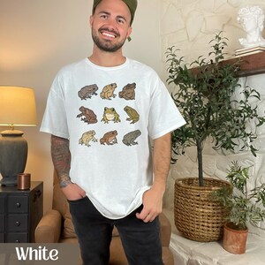 May include: White t-shirt with a graphic print of nine brown and green toads. The toads are arranged in three rows of three. The shirt is being worn by a person with a light brown beard and a green baseball cap.