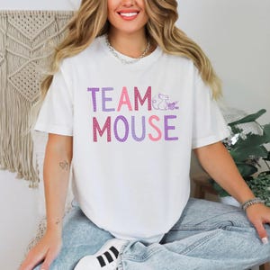 May include: White t-shirt with the text "TEAM MOUSE" in pink and purple letters. The text is outlined in white dots. A small purple mouse graphic is next to the word "TEAM".