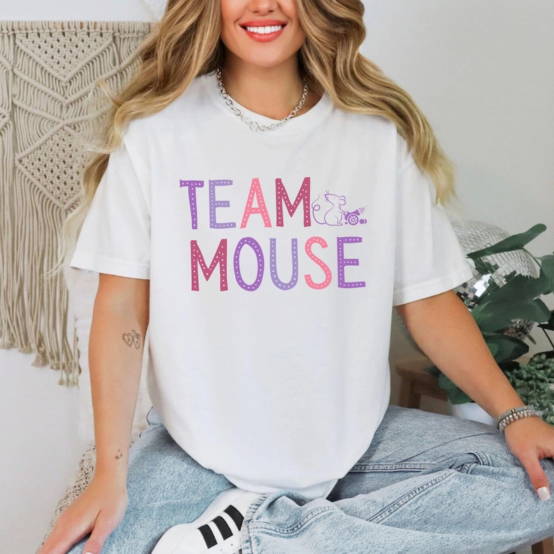 May include: White t-shirt with the text "TEAM MOUSE" in pink and purple letters. The text is outlined in white dots. A small purple mouse graphic is next to the word "TEAM".