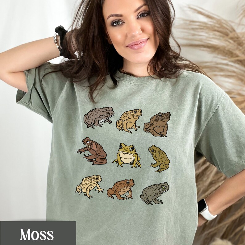 May include: A light green t-shirt with a graphic print of nine brown, green, and yellow frogs. The shirt has the word "Moss" printed on the bottom.