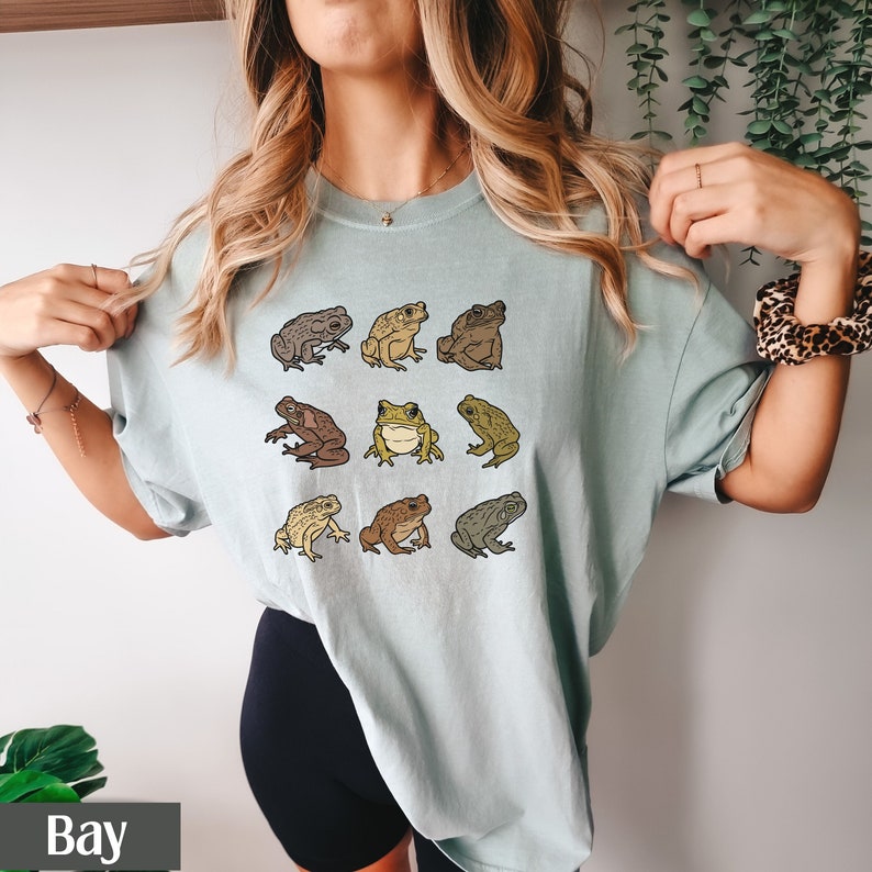 May include: A light blue t-shirt with a graphic of nine brown and green frogs. The frogs are arranged in a grid pattern on the shirt.