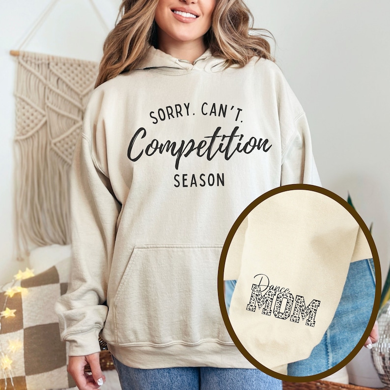 May include: A beige hoodie with the text "Sorry. Can't. Competition Season" printed on the front. The hoodie has a pocket on the front and a hood. A close-up of the sleeve shows the text "Dance Mom" printed in black with a leopard print pattern.