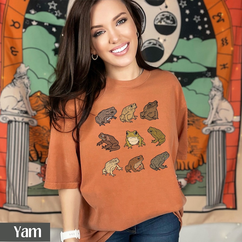 May include: A woman wearing an orange t-shirt with a graphic of nine different frogs in various poses. The shirt has a crew neck and short sleeves.