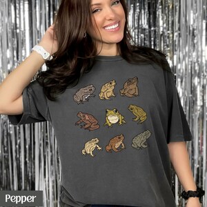 May include: A dark gray t-shirt with a vintage look featuring a pattern of nine different frogs in various shades of brown, green, and yellow. The frogs are arranged in a grid pattern on the shirt.