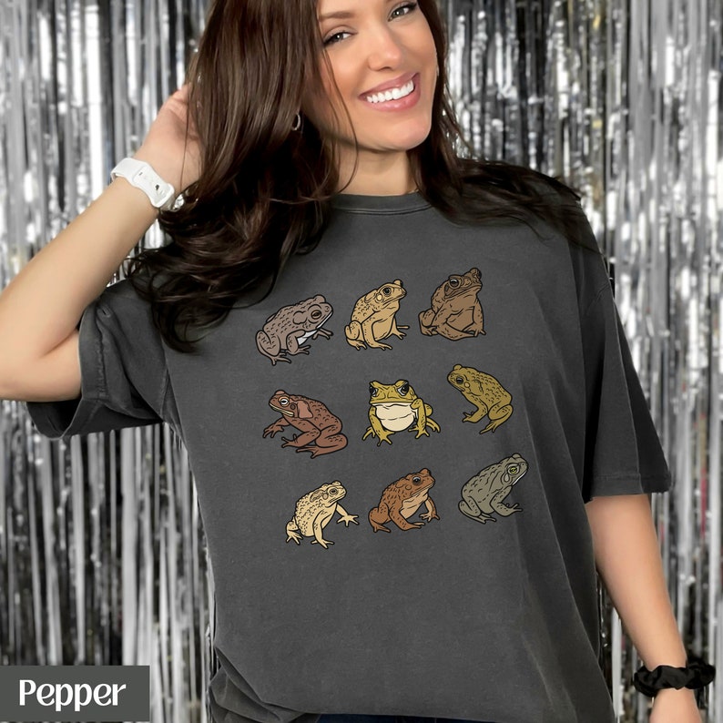 May include: A dark gray t-shirt with a vintage look featuring a pattern of nine different frogs in various shades of brown, green, and yellow. The frogs are arranged in a grid pattern on the shirt.
