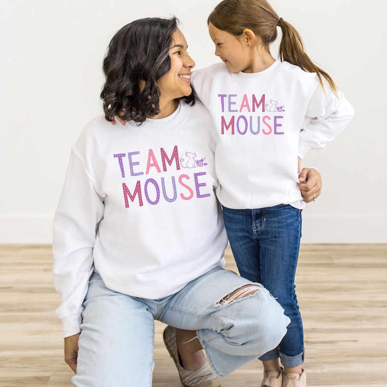 May include: Two people wearing white sweatshirts with the text "Team Mouse" in pink and purple lettering. The sweatshirts feature a small mouse graphic. The person in the front is kneeling and the person in the back is standing.