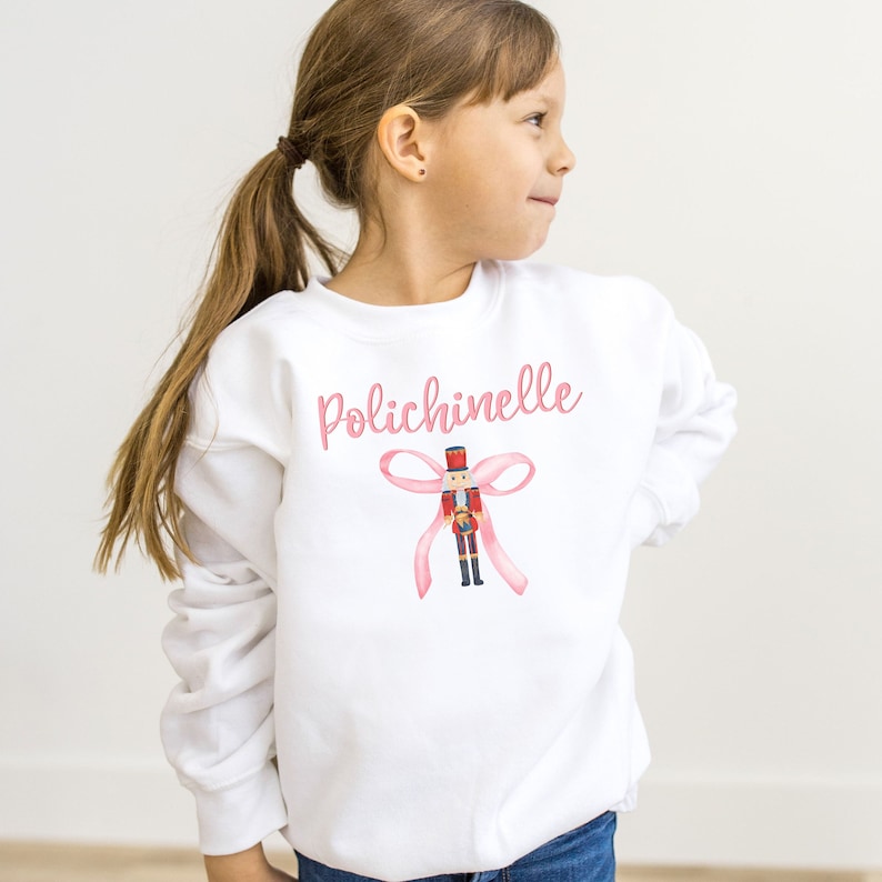 Girl in a white Gildan brand crewnecked sweatshirt.  The chest reads "Polichinelle" in pink cursive above a coquette-style bow with a watercolor Nutcracker.  Sweatshirts in youth sizes in White, Maroon, Forest Green, and Dark Grey Heather.