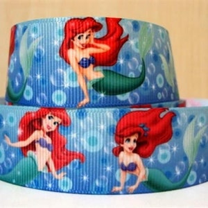 May include: Two stacked rolls of light blue ribbon featuring a repeating pattern of a mermaid with red hair and a green tail. The mermaid is wearing a purple seashell bra. The background has white bubbles and vertical lines.