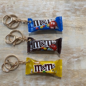 May include: Three keychains featuring miniature M&M's candy bar replicas. The keychains have gold-colored hardware. One candy bar is blue, one is brown, and one is yellow. Each candy bar has the M&M's logo and a cartoon character.