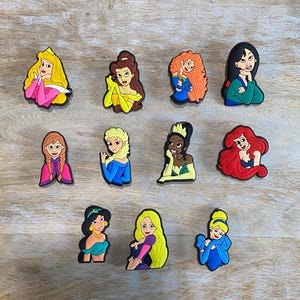 May include: A collection of colorful cartoon character shoe charms. The charms feature various princesses with distinct hairstyles and outfits, including blonde, red, and black hair, and dresses in shades of pink, yellow, blue, and green. The charms are arranged on a light brown wooden surface.
