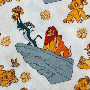May include: Fabric featuring characters from The Lion King. The design includes Simba, Mufasa, and Nala on a light blue background. Other elements include paw prints and butterflies. The fabric is suitable for various crafting projects.