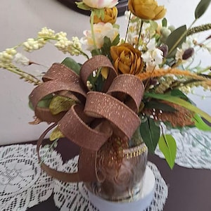 May include: A floral arrangement featuring artificial mustard yellow and cream-colored flowers, green foliage, and tall grass-like stems. A large, shimmering bronze-colored ribbon with a glittery texture is tied around the base of the clear glass vase.