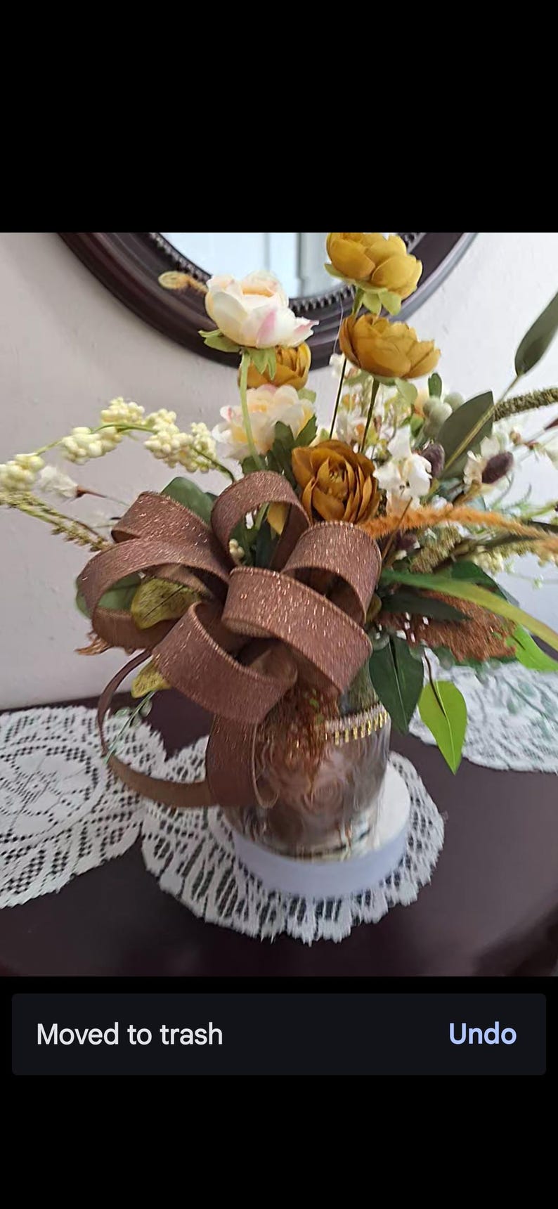 May include: A floral arrangement featuring artificial mustard yellow and cream-colored flowers, green foliage, and tall grass-like stems. A large, shimmering bronze-colored ribbon with a glittery texture is tied around the base of the clear glass vase.
