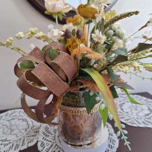 May include: A decorative floral arrangement in a glass vase featuring mustard yellow and cream-colored artificial flowers, green foliage, and small white berries. A large, glittery bronze-colored ribbon is tied to the side of the vase.