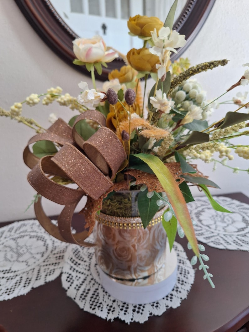 May include: A decorative floral arrangement in a glass vase featuring mustard yellow and cream-colored artificial flowers, green foliage, and small white berries. A large, glittery bronze-colored ribbon is tied to the side of the vase.