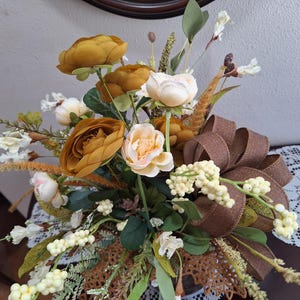 May include: A floral arrangement featuring mustard yellow and cream-colored faux flowers, green foliage, and tall wheat-like stalks. A large, textured brown ribbon bow is integrated into the bouquet, which is accented with small white berries.