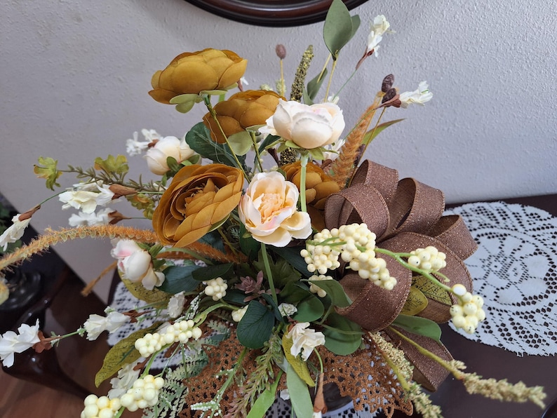 May include: A floral arrangement featuring mustard yellow and cream-colored faux flowers, green foliage, and tall wheat-like stalks. A large, textured brown ribbon bow is integrated into the bouquet, which is accented with small white berries.
