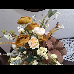 May include: A floral arrangement featuring mustard yellow and cream-colored roses, white berry clusters, and green foliage. A large, textured brown ribbon with a subtle shimmer is integrated into the bouquet, which is accented by tall, thin grasses.