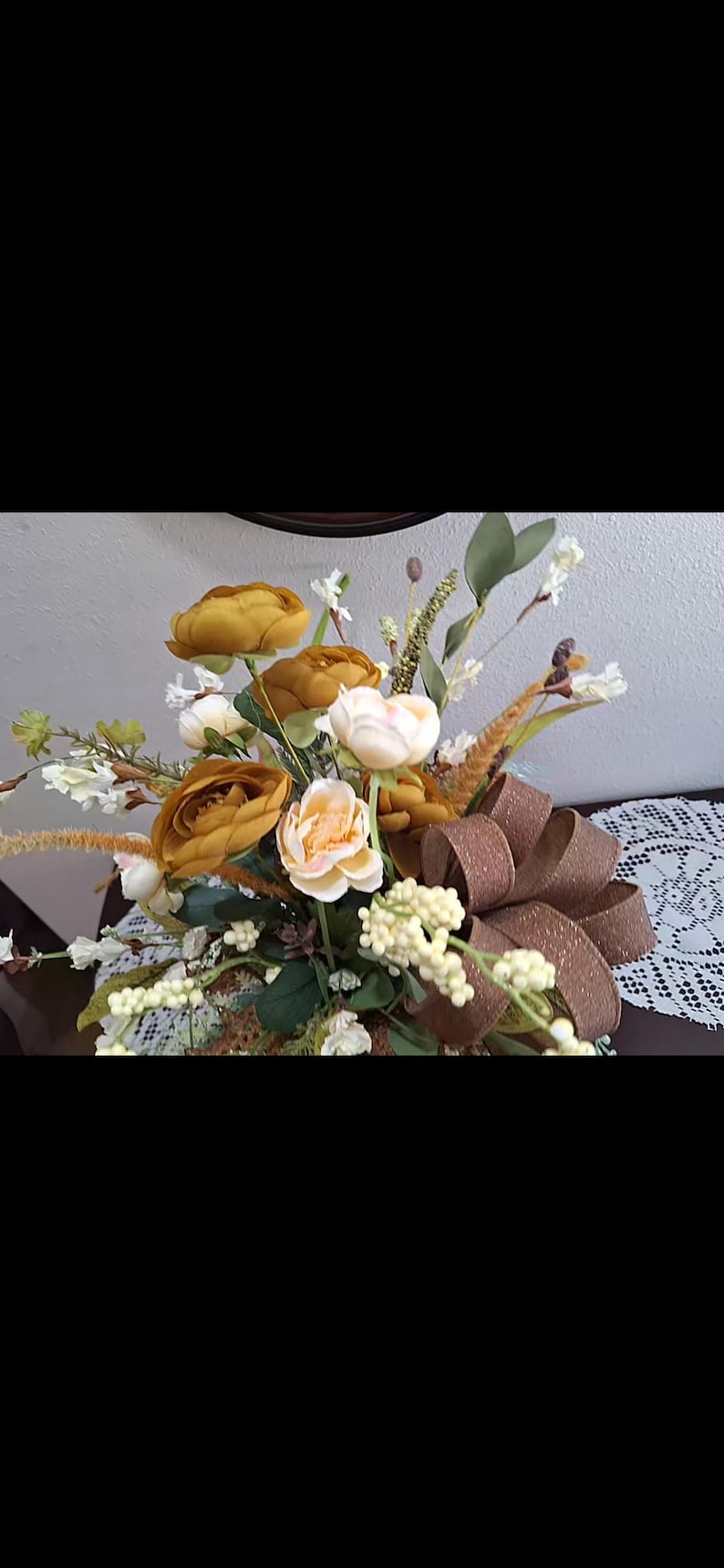 May include: A floral arrangement featuring mustard yellow and cream-colored roses, white berry clusters, and green foliage. A large, textured brown ribbon with a subtle shimmer is integrated into the bouquet, which is accented by tall, thin grasses.