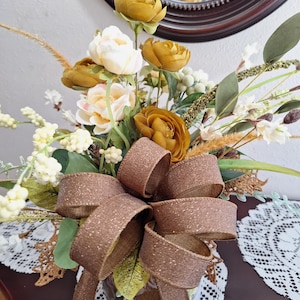 May include: A floral arrangement featuring mustard yellow and cream-colored artificial flowers, green foliage, and white berry sprigs. A large, glittery brown burlap bow is tied at the front, adding texture to the bouquet of mixed botanical elements.