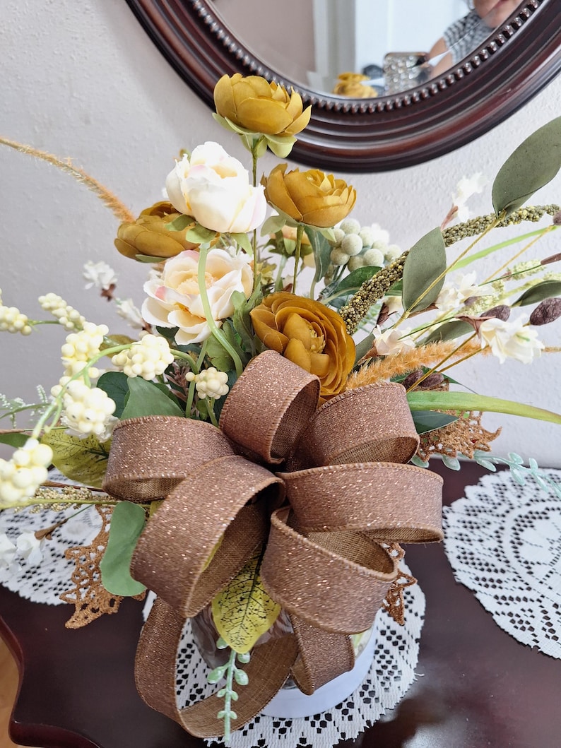 May include: A floral arrangement featuring mustard yellow and cream-colored artificial flowers, green foliage, and white berry sprigs. A large, glittery brown burlap bow is tied at the front, adding texture to the bouquet of mixed botanical elements.
