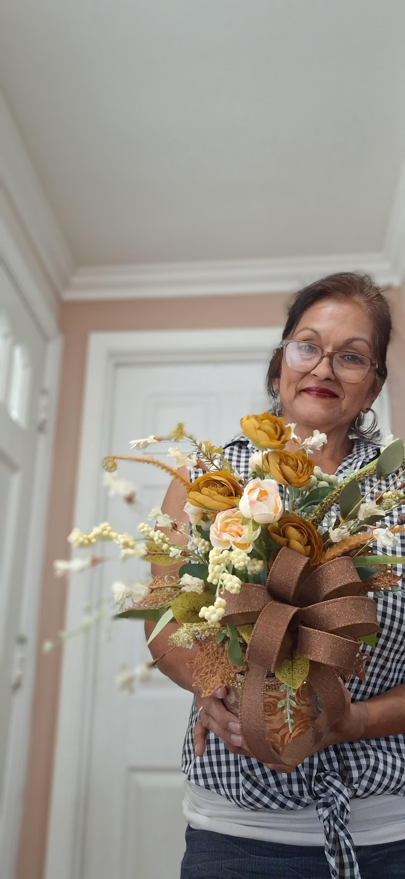 May include: A decorative floral arrangement featuring mustard yellow and cream-colored artificial roses, white berries, and green foliage. The bouquet is accented with a large, textured brown ribbon bow tied around the base of the container.