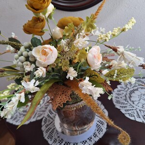 May include: A floral arrangement featuring mustard yellow and pale pink flowers, green foliage, and small white berries. The bouquet is housed in a brown, textured vase with a decorative band, accented by a brown lace leaf-shaped embellishment.