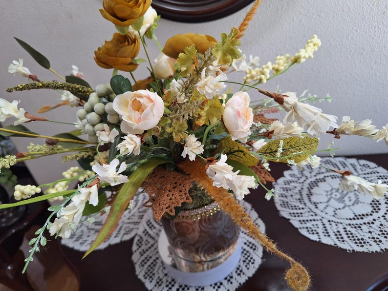 May include: A floral arrangement featuring mustard yellow and pale pink flowers, green foliage, and small white berries. The bouquet is housed in a brown, textured vase with a decorative band, accented by a brown lace leaf-shaped embellishment.
