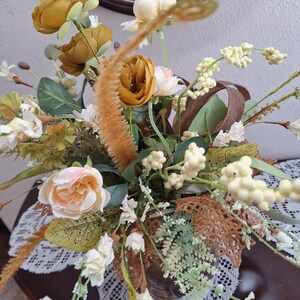 May include: A floral arrangement featuring mustard yellow and pale pink roses, white berry clusters, green foliage, and brown textured leaves. The bouquet includes tall, fuzzy, orange-toned stalks and delicate white blossoms on thin stems.