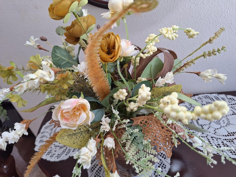 May include: A floral arrangement featuring mustard yellow and pale pink roses, white berry clusters, green foliage, and brown textured leaves. The bouquet includes tall, fuzzy, orange-toned stalks and delicate white blossoms on thin stems.