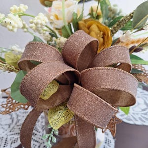 May include: A decorative floral arrangement featuring mustard yellow and cream-colored flowers, green foliage, and white berries. A large, shimmering bronze-colored ribbon bow with a glittery texture is tied prominently in the center of the bouquet.