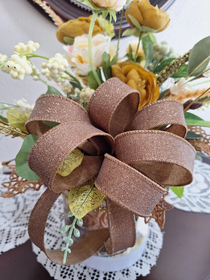May include: A decorative floral arrangement featuring mustard yellow and cream-colored flowers, green foliage, and white berries. A large, shimmering bronze-colored ribbon bow with a glittery texture is tied prominently in the center of the bouquet.