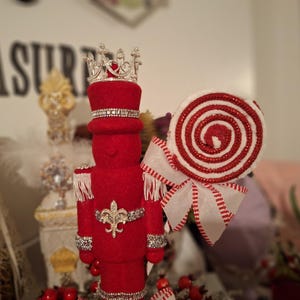 May include: A red felt nutcracker figurine wearing a silver crown and a silver and red striped hat. The nutcracker is holding a red and white candy cane.