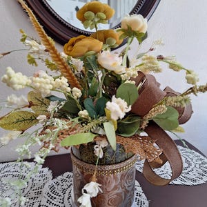 May include: A floral arrangement in a cylindrical glass vase featuring mustard yellow and cream-colored artificial flowers, green foliage, and tall wheat-like stems. The vase is wrapped with a brown ribbon and a decorative gold-toned trim.