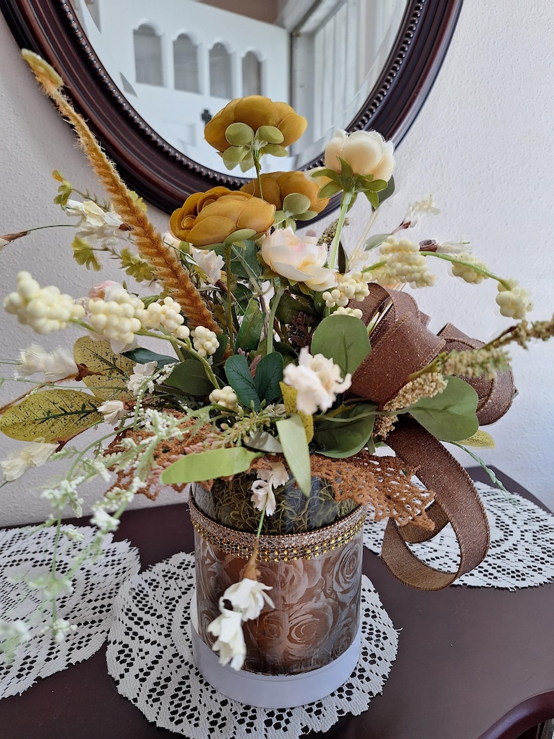 May include: A floral arrangement in a cylindrical glass vase featuring mustard yellow and cream-colored artificial flowers, green foliage, and tall wheat-like stems. The vase is wrapped with a brown ribbon and a decorative gold-toned trim.
