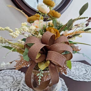 May include: A floral arrangement featuring mustard yellow and cream-colored artificial flowers, green foliage, and white berries. A large, glittery brown ribbon bow is tied around the center of the glass vase, which is decorated with gold trim.