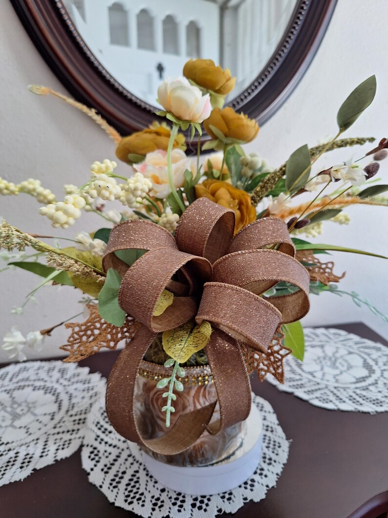 May include: A floral arrangement featuring mustard yellow and cream-colored artificial flowers, green foliage, and white berries. A large, glittery brown ribbon bow is tied around the center of the glass vase, which is decorated with gold trim.