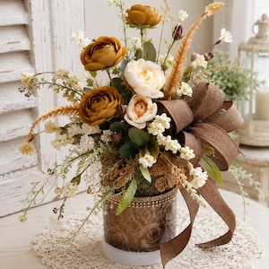 May include: A floral arrangement featuring mustard yellow and cream-colored faux flowers, green foliage, and tall grasses. The bouquet is housed in a cylindrical glass vase with a rose pattern, accented by a large, glittery brown fabric bow.
