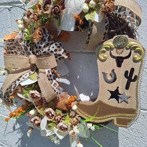 May include: A decorative wreath featuring a grapevine base, artificial brown and white roses, orange berries, and leopard print burlap ribbons. A wooden cowboy boot cutout is attached, decorated with a bull skull, horseshoe, cactus, and star.