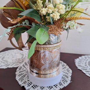 May include: A cylindrical vase featuring a metallic rose pattern and a gold-toned beaded trim at the top rim. The vase is filled with an arrangement of artificial greenery, white berries, and brown textured leaves with a brown ribbon accent.