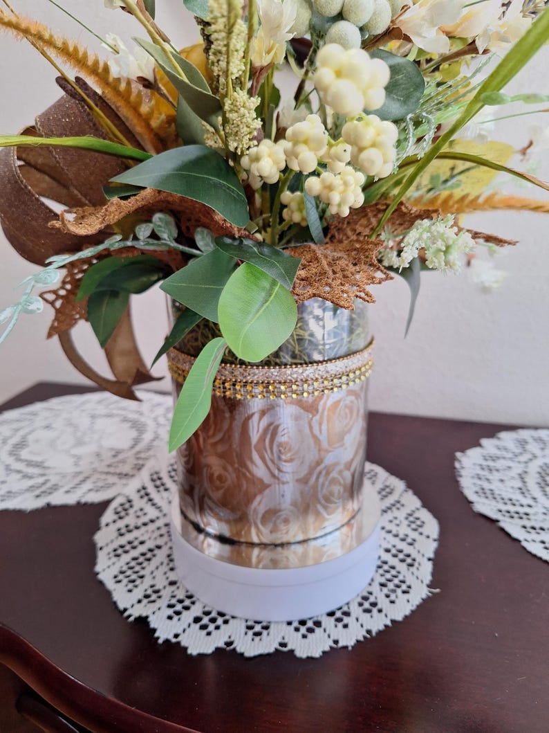 May include: A cylindrical vase featuring a metallic rose pattern and a gold-toned beaded trim at the top rim. The vase is filled with an arrangement of artificial greenery, white berries, and brown textured leaves with a brown ribbon accent.