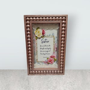 May include: A framed decorative wall art piece with a rose floral design and the text "Sister She is Clothed with Strength and Dignity She can Laugh at The Days to Come Proverbs 31:25". The frame is rose gold with a beaded border.