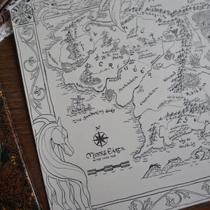 May include: A black and white line art map of Middle-earth in the Third Age printed on paper. The map features intricate borders with dragon and horse illustrations, a compass rose, a scale bar, and text labels for regions like The Shire.