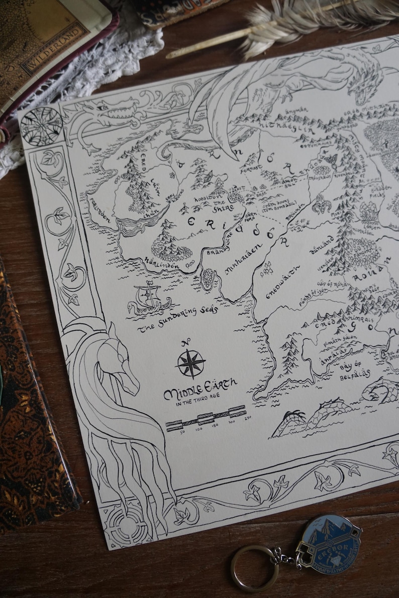 May include: A black and white line art map of Middle-earth in the Third Age printed on paper. The map features intricate borders with dragon and horse illustrations, a compass rose, a scale bar, and text labels for regions like The Shire.