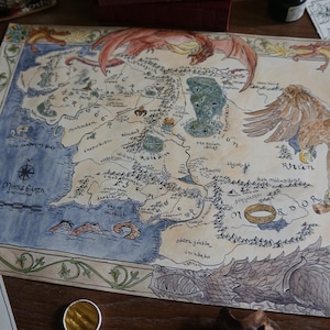 May include: A hand-painted map of Middle-earth from The Lord of the Rings. The map features the land of Mordor, the Shire, and the Misty Mountains. The map is surrounded by a decorative border.