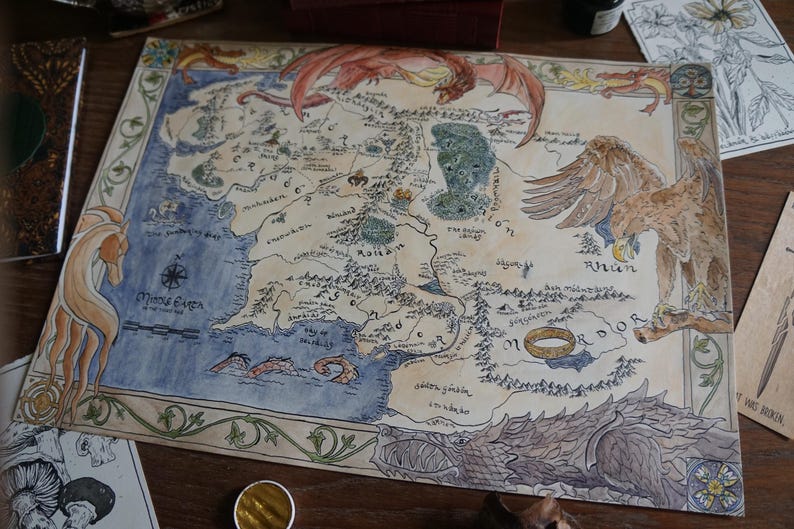 May include: A hand-drawn map of Middle-earth on parchment-colored paper, featuring intricate ink illustrations of a dragon, a large eagle, and the One Ring. The map includes detailed geographical labels, a compass rose, and decorative borders.