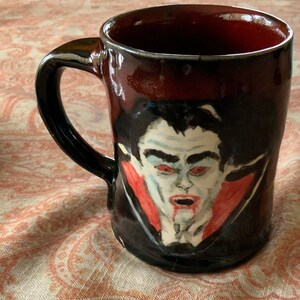 Peut inclure: Une tasse en céramique noire avec un bord rouge et une image peinte à la main d'un vampire aux yeux rouges et à la cape rouge. La tasse est sur une surface en tissu à motifs.