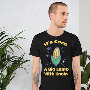 以下が含まれることがあります： 白いコーンと黄色のコーンのグラフィックと「It's Corn. A Big Lump With Knobs」というテキストが書かれた黒いTシャツです。