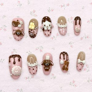 May include: A set of ten artificial nails with a pink base and various decorative elements. Designs include chocolate drips, teddy bears, bows, and bunny figures. Some nails feature white polka dots and a chocolate bar.
