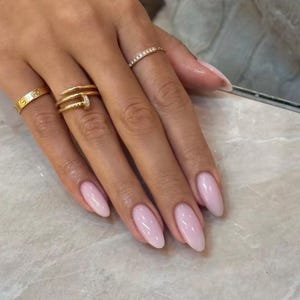Könnte beinhalten: Eine Hand mit mandelförmigen Nägeln, die in einem glänzenden Hellrosa lackiert sind. Die Finger sind mit drei Goldringen geschmückt: einem schlichten Ring am Zeigefinger, einem doppelten Nagelring am Mittelfinger und einem dünnen Ring am Ringfinger.